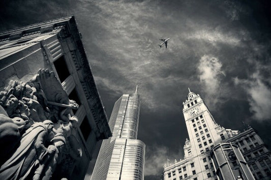 This stunning black and white photograph captures the iconic Chicago skyline with a unique perspective looking up at the Wrigley Building and Trump Tower. The image also includes a plane soaring through the space between the two buildings, adding a dynamic and dramatic touch to the composition. A perfect piece of art for anyone who loves Chicago architecture and cityscapes.